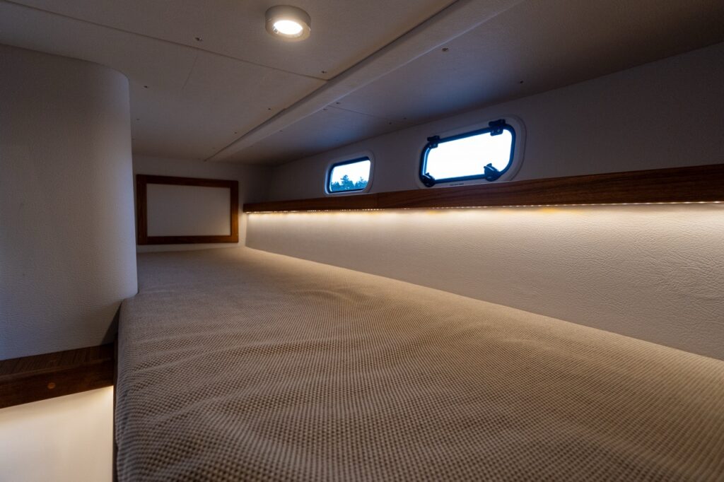 Single aft berth on the Targa 32 with a long upholstered sleeping surface, hull-side portlights, teak trim, and warm indirect LED lighting along the berth edge.