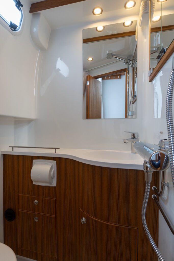 Head compartment vanity on the Targa 32 featuring a molded white sink, teak cabinetry, mirror with overhead lighting, and integrated shower fixture.