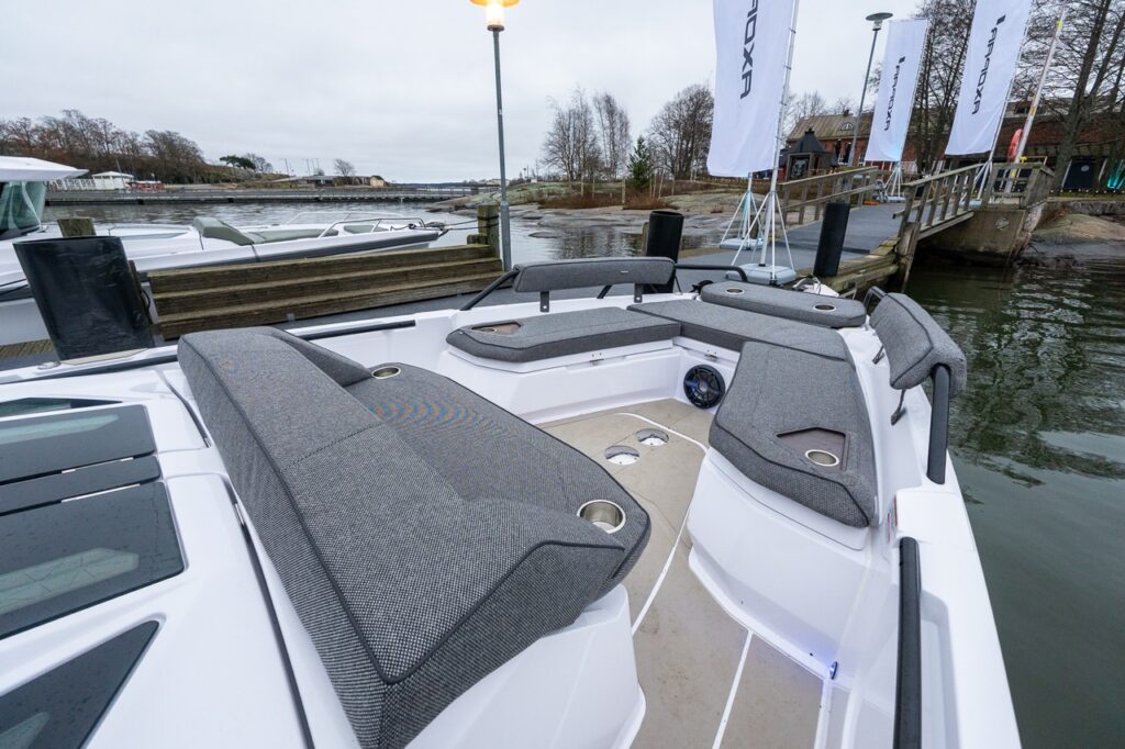 Angled view of the Axopar 38 bow seating area, showing modular gray upholstered lounges, integrated cupholders, armrests, and an open social layout at the dock.