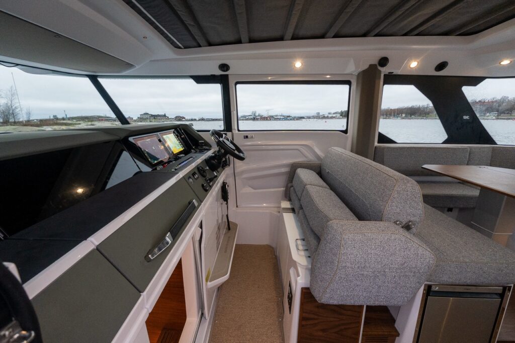Interior view of the Axopar 38 pilothouse showing the bench-style helm seat alongside the helm, dual Multi Function Displays (MFDs), wide side windows, and the aft dining area visible behind the helm.