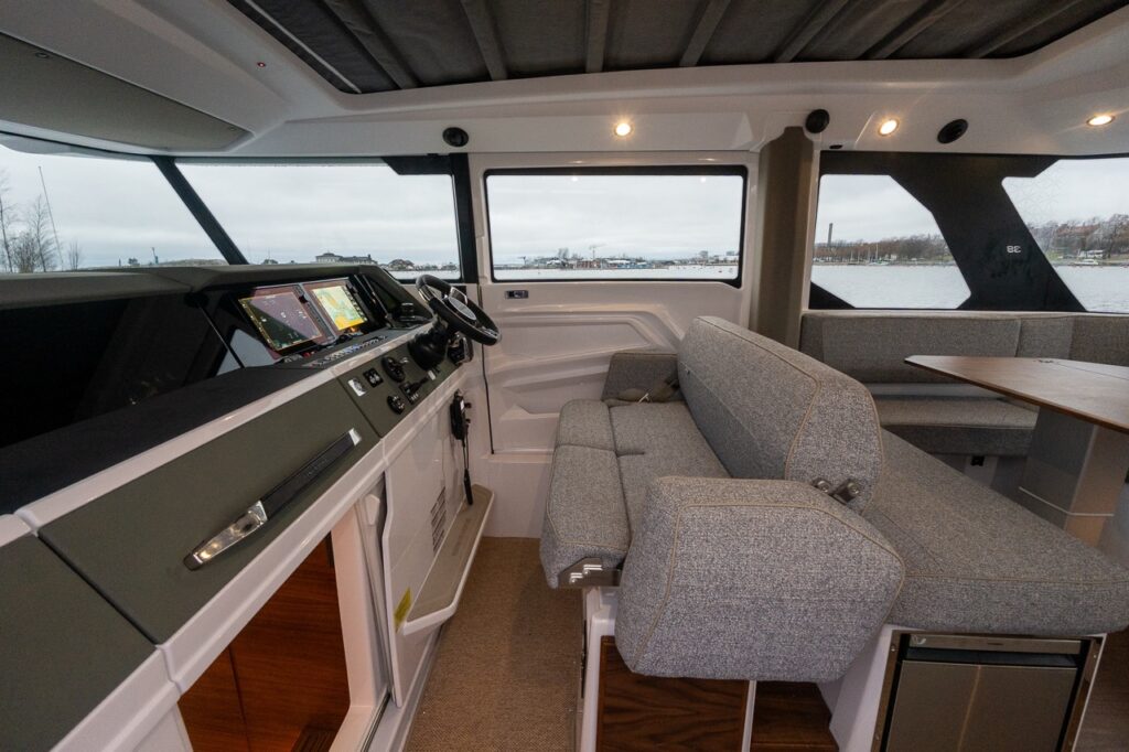 Interior view of the Axopar 38 pilothouse showing the bench-style helm seat positioned beside the helm, with dual Multi Function Displays (MFDs), wide side windows, and clear sightlines throughout the cabin.
