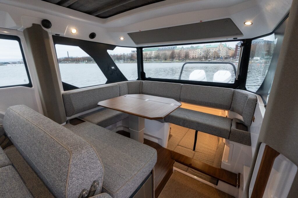 Aft section of the Axopar 38 pilothouse showing wraparound lounge seating arranged around a central table, panoramic side and aft windows, overhead lighting, and integrated access down to the aft cabin.
