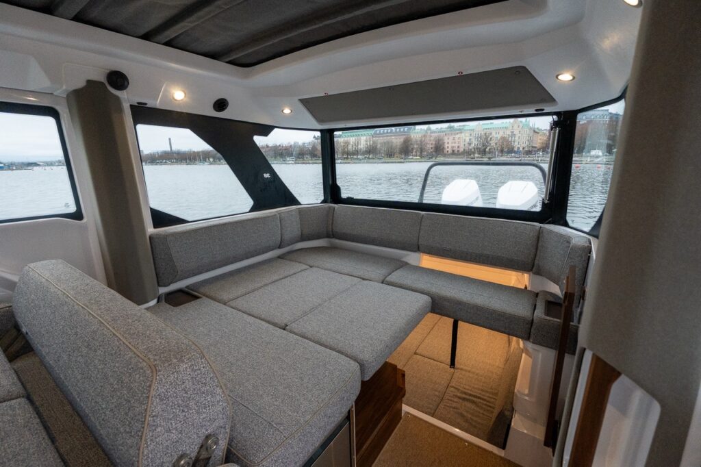 Aft section of the Axopar 38 pilothouse showing wraparound lounge seating converted into a large berth, panoramic side and aft windows, overhead lighting, and integrated access down to the aft cabin.