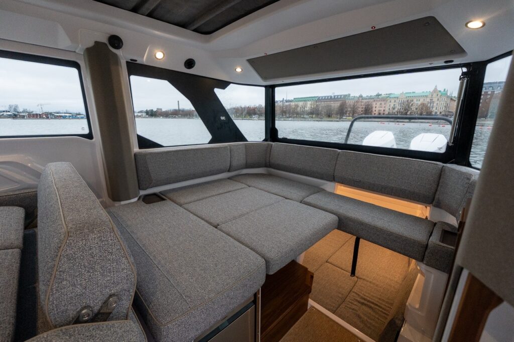 Aft seating area inside the Axopar 38 pilothouse showing wraparound lounge seating converted into a large berth, with panoramic windows, overhead lighting, and integrated access down to the aft cabin.