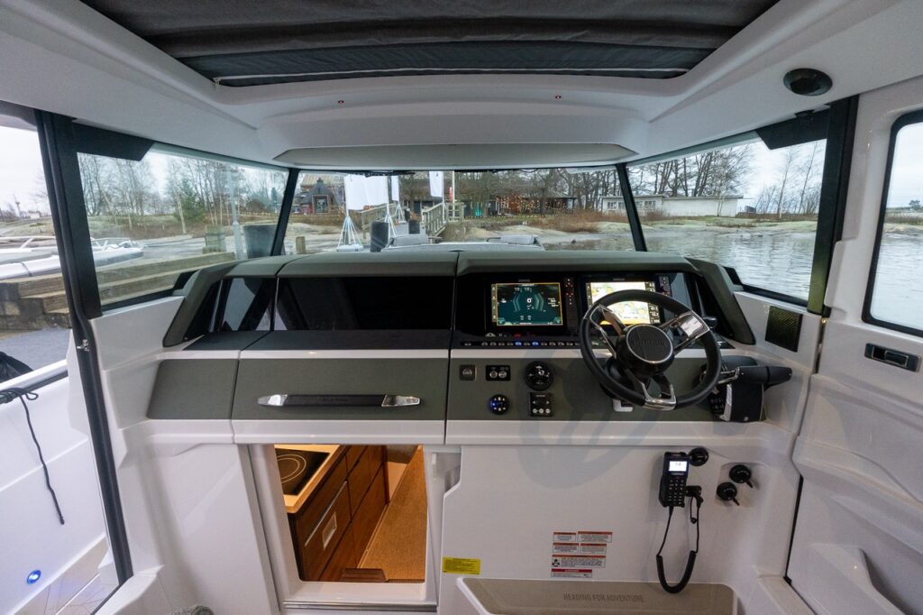 Wide interior view of the Axopar 38 pilothouse showing the full helm layout with dual Simrad Multi Function Displays (MFDs), BRABUS steering wheel, Mercury controls, panoramic windshield, and the stairway leading down to the forward cabin.