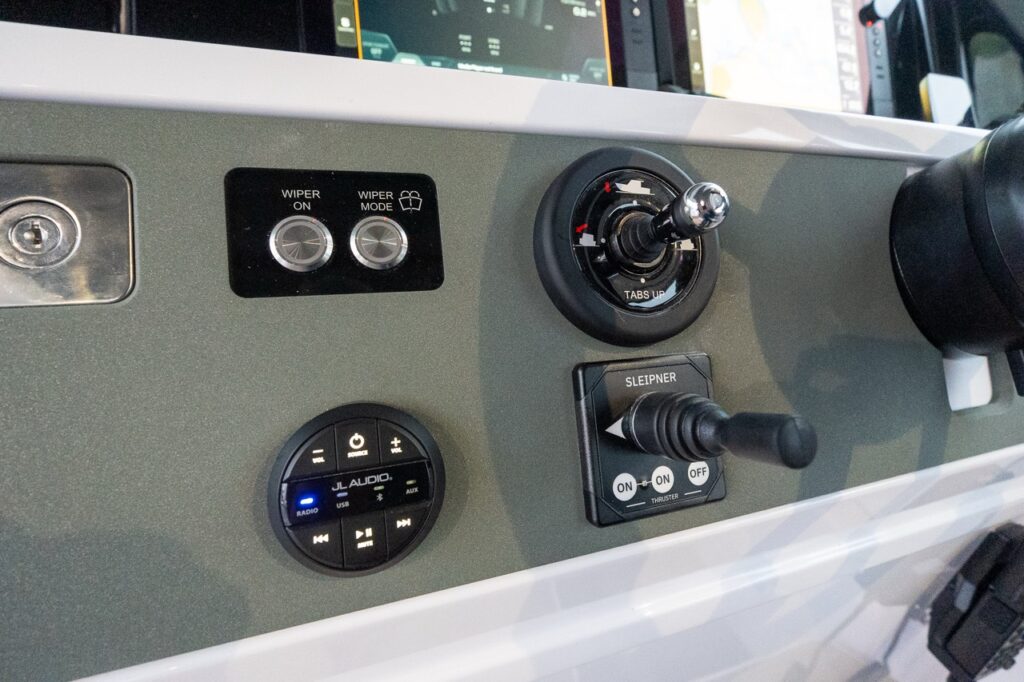 Close-up of the Axopar 38 helm controls showing windshield wiper switches, JL Audio stereo control, trim tab joystick, and Sleipner bow thruster control integrated into a clean, modern dashboard.