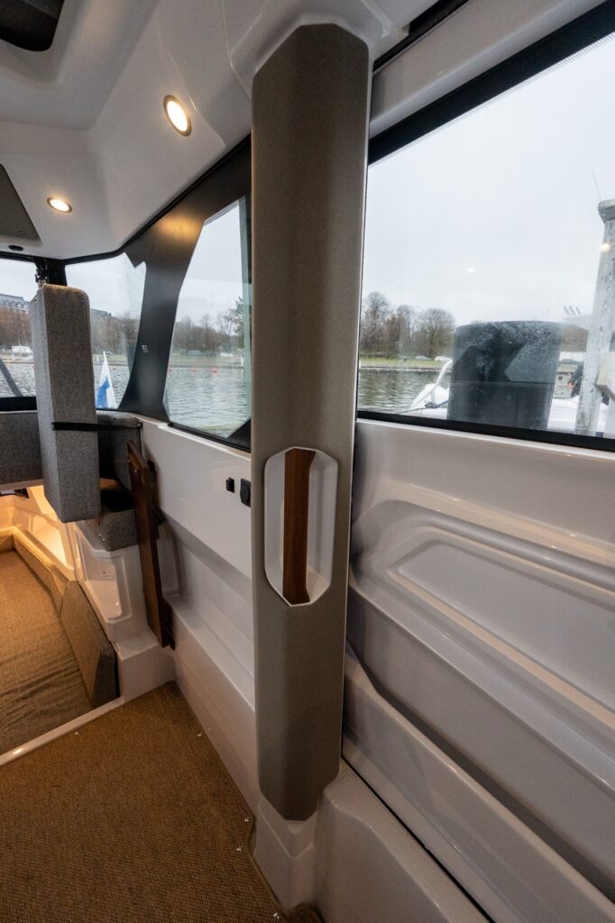 Interior detail of the all-new Axopar 38 pilothouse showing a padded vertical grab handle integrated into the side pillar, surrounded by panoramic windows, modern upholstery, and clean Scandinavian interior finishes.