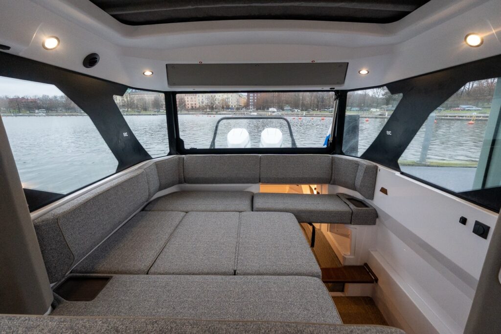 Interior view of the Axopar 38 pilothouse aft seating area showing wraparound lounge seating that converts into a large berth, panoramic windows, and the new opening aft window connecting the pilothouse to the aft deck with water views outside.
