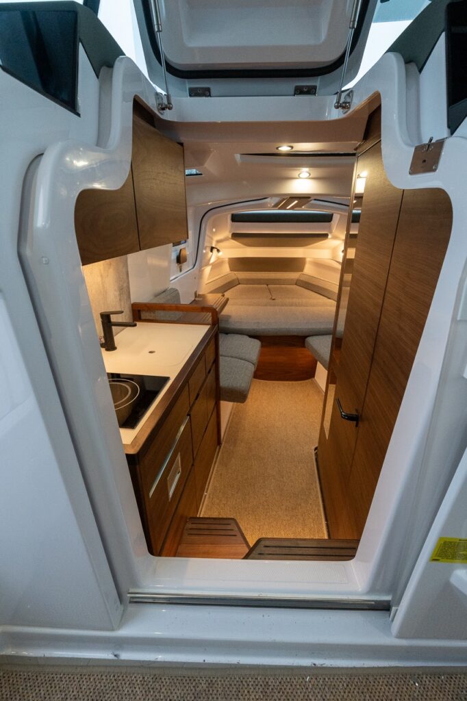 View into the forward cabin of the all-new Axopar 38 showing the entryway with steps down, a compact galley to port, a separate enclosed head compartment to starboard, and a forward seating area that converts into a berth, finished with warm wood cabinetry and modern lighting.