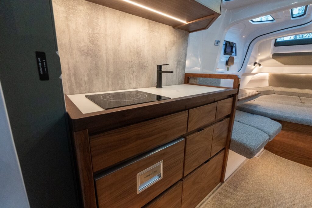 Axopar 38 forward cabin galley with induction cooktop, integrated sink, Dometic refrigerator, warm Nordic wood cabinetry, and LED accent lighting.