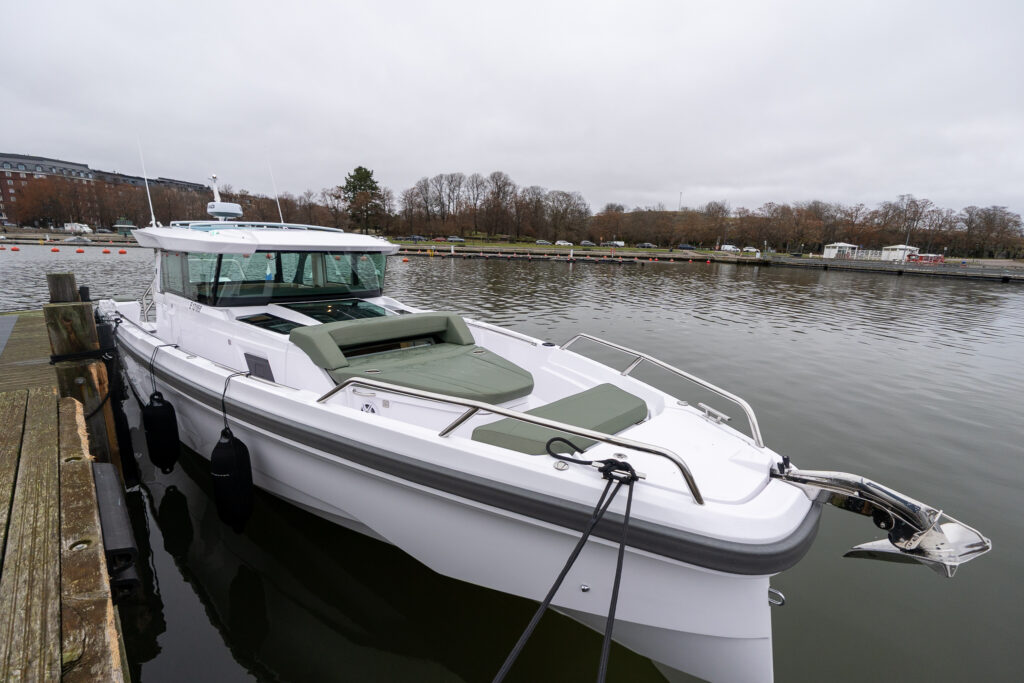 Axopar 38 Cross Cabin moored at the dock, shown from the bow quarter with clean white hull lines, enclosed pilothouse, forward sunpad, stainless bow rail, and calm harbor surroundings.